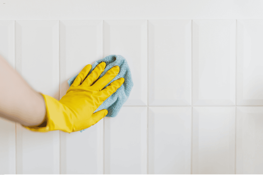 Mão com luva amarela limpando azulejos brancos com pano de microfibra.