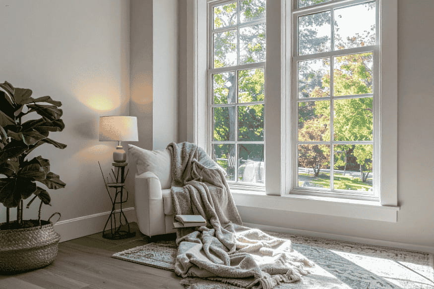 Sala de estar minimalista com poltrona branca, manta, luminária e janela ampla com vista para jardim.