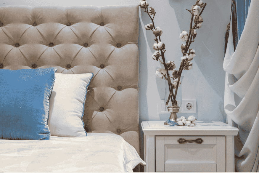 Cabeceira estofada bege com capitonê em quarto decorado com mesa de cabeceira.