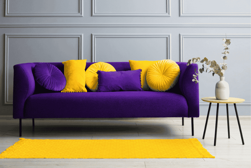 Sala moderna com sofá roxo e almofadas amarelas combinando com tapete amarelo, criando contraste harmonioso na decoração.