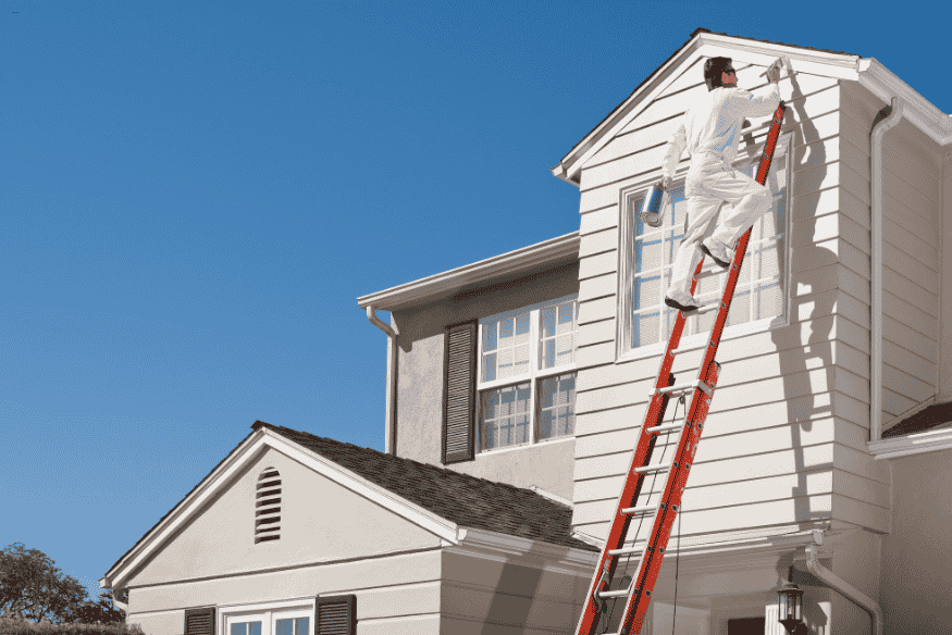 Pintor em escada alta aplicando tinta branca na parede externa da casa.