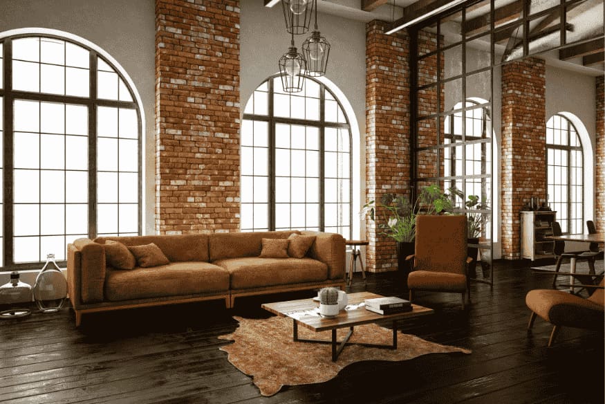 Loft industrial com sofá marrom, parede de tijolos e janelas grandes de estrutura metálica.