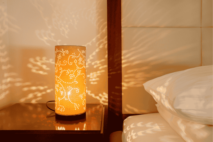 Luminária de mesa cilíndrica perfurada emitindo luz suave e criando padrões na parede.