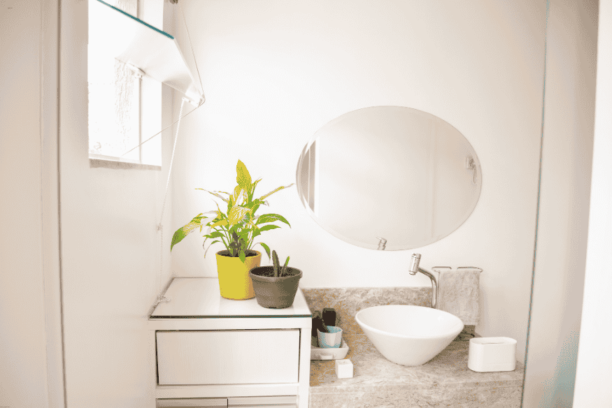 Banheiro claro com espelho redondo e plantas decorativas iluminado por luz natural.
