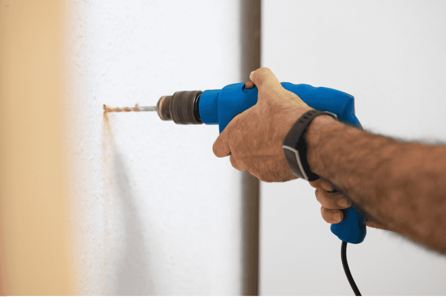 Mão usando furadeira azul para perfurar parede antes de instalar prateleira.