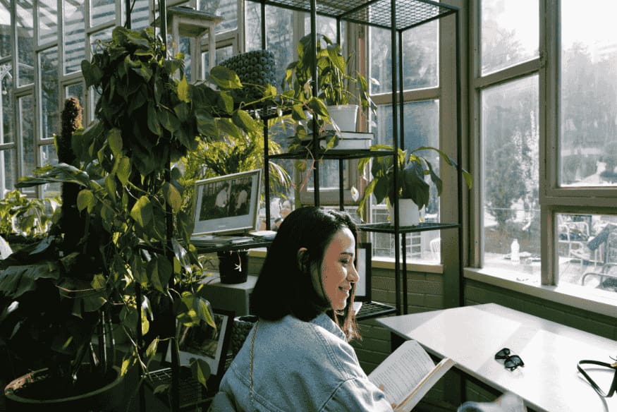 Mulher lendo em espaço com prateleiras repletas de plantas junto a janelas amplas.