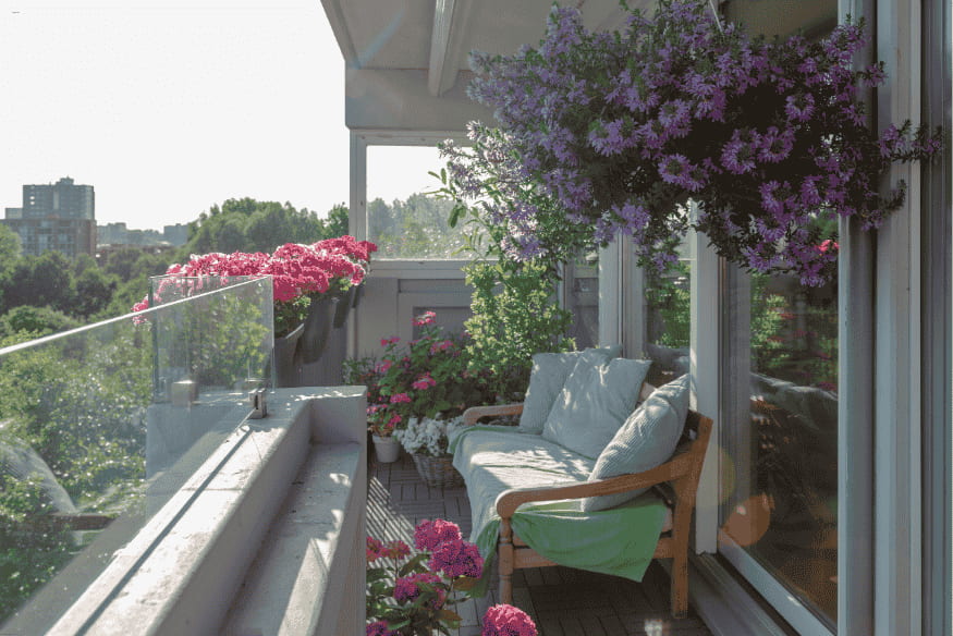 Varanda decorada com flores e folhagens, sofá de madeira e vista para área verde.
